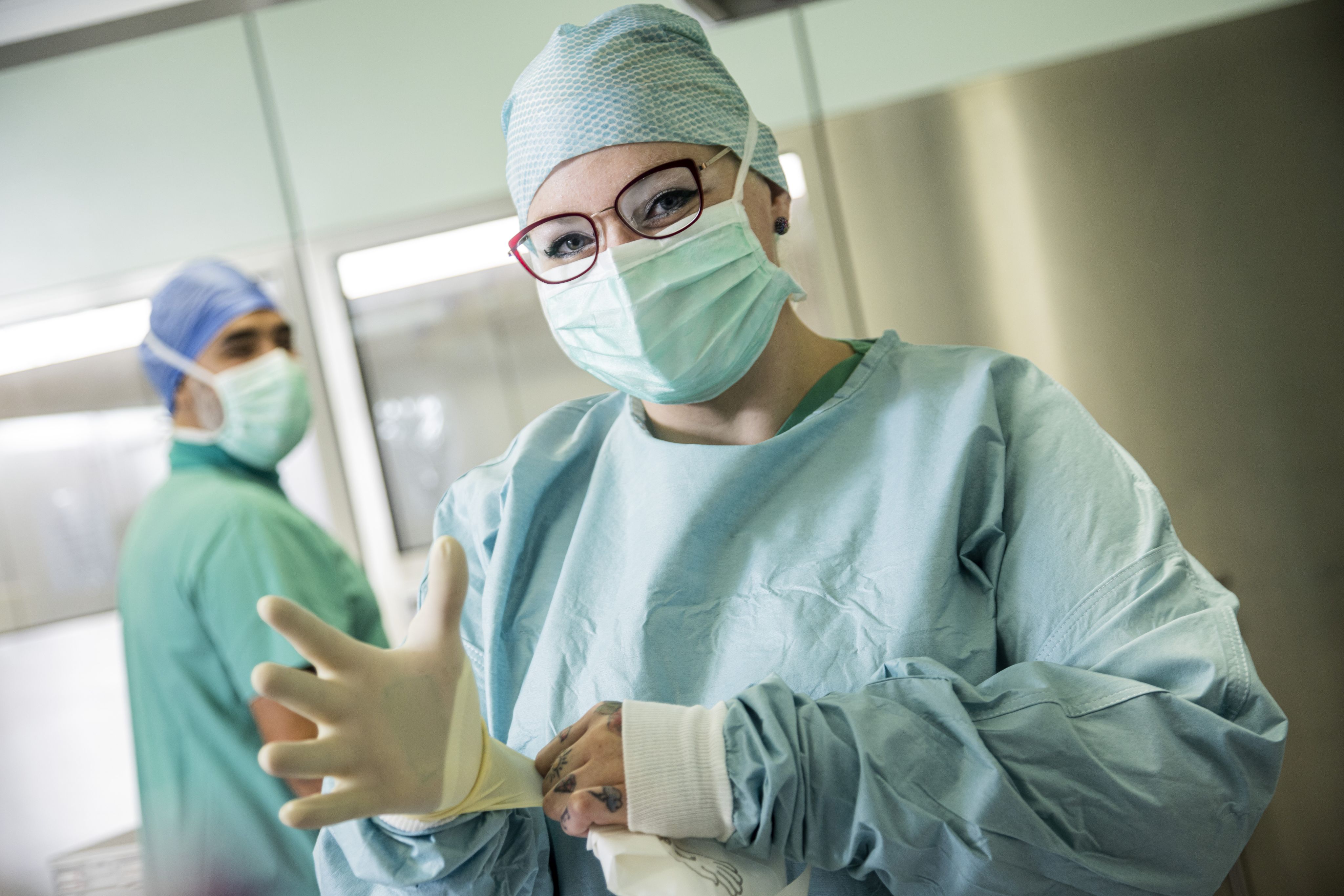 ine Person in OP-Kleidung steht in einem Operationssaal. Sie trägt einen Mund-Nasen-Schutz, eine OP-Haube und medizinische Handschuhe. Im Hintergrund ist eine zweite Person zu sehen.
