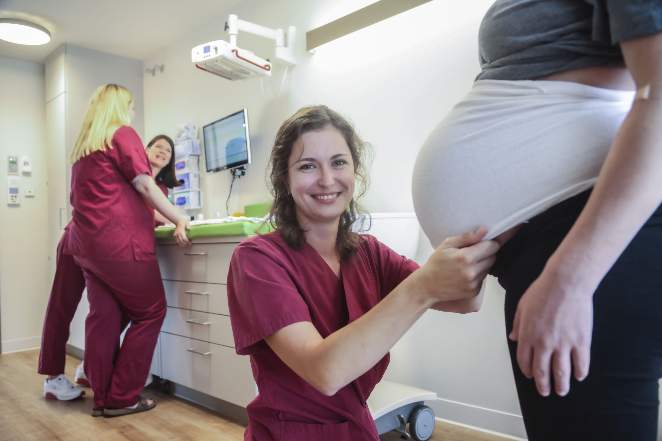 Hebamme steht vor dem Bauch einer schwangeren Person. Im Hintergrund sind weitere Hebammen in beerenfarbener Berufskleidung zu erkennen. 