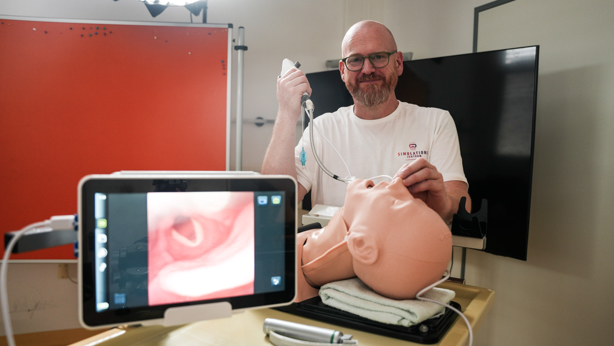 Männliche Person führt eine Inkubation an einem Simulationskopf durch. Im Vordergrund steht ein kleiner Bildschirm mit einem Bild der Luftröhre. 
