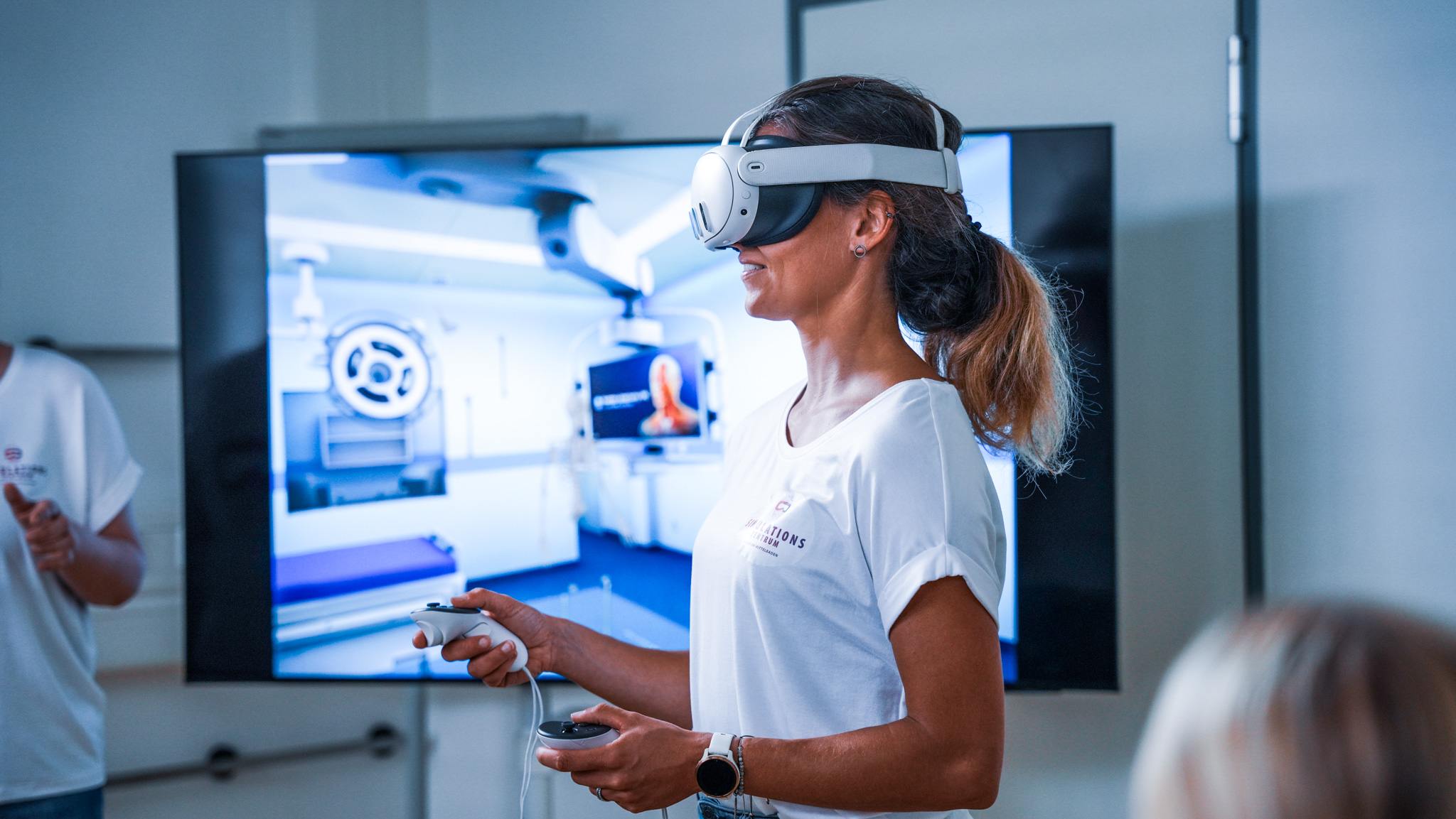 Weibliche Person hat eine VR Brille an und zwei Cursor in der Hand. Im Hintergrund ist ein Fernseher mit einem OP Szenario.