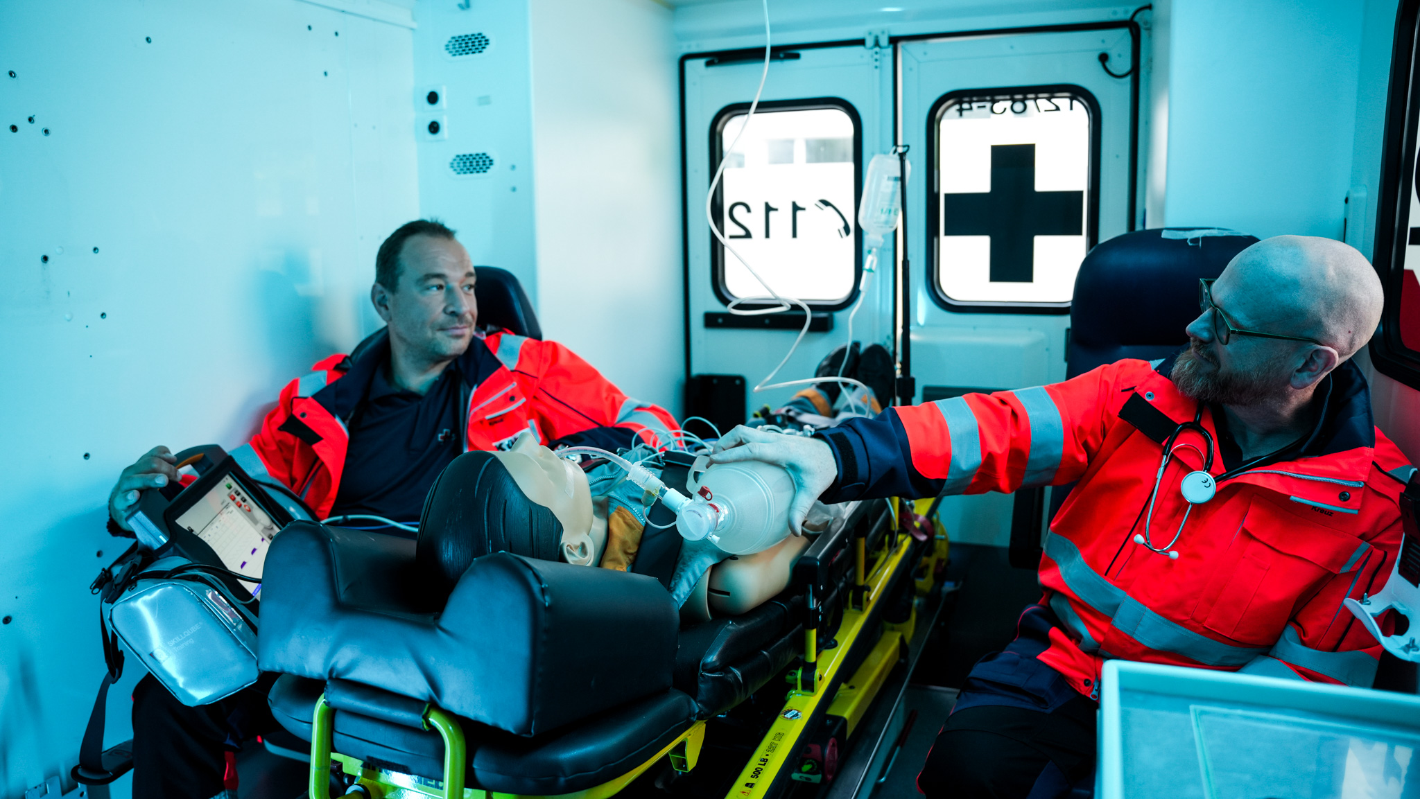 zwei Personen sitzen mit orangener Kleidung vom Rettungsdienst im hinteren Teil eines Rettungswagens. Das Bild hat einen bläulichen Farbstich