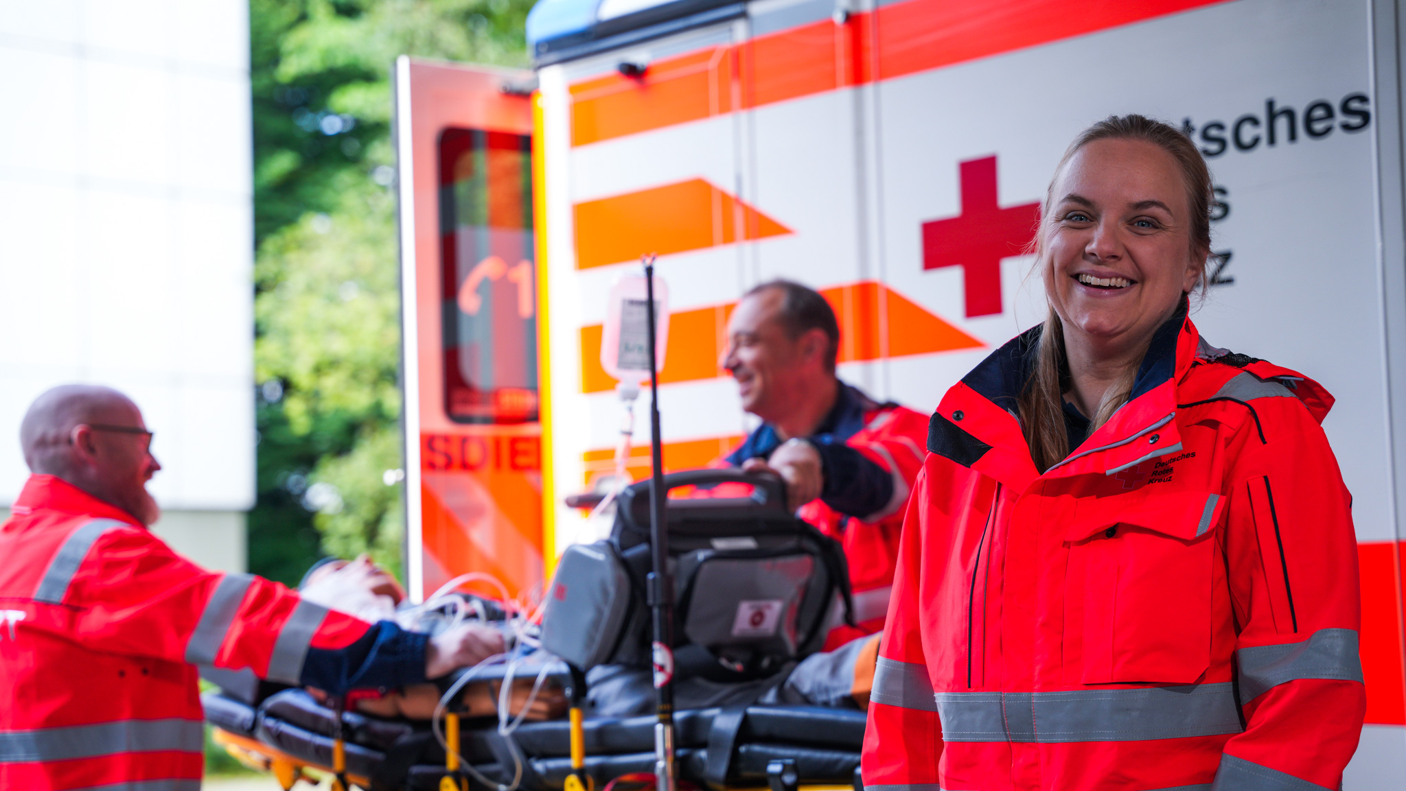 Weibliche Person steht in orangener Kleidung seitlich vor einem Rettungswagen und schaut in die Kamera. im Hintergrund sind zwei weitere Rettungssanitäter mit Trage. 