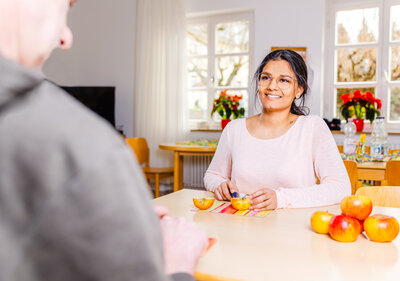 Person schneidet einen Apfel für eine gegenübersitzende Person. Die Person im Gegenüber ist nur von hinten zu sehen,