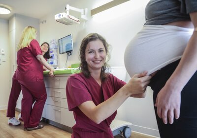 Hebamme steht vor dem Bauch einer schwangeren Person. Im Hintergrund sind weitere Hebammen in beerenfarbener Berufskleidung zu erkennen. 