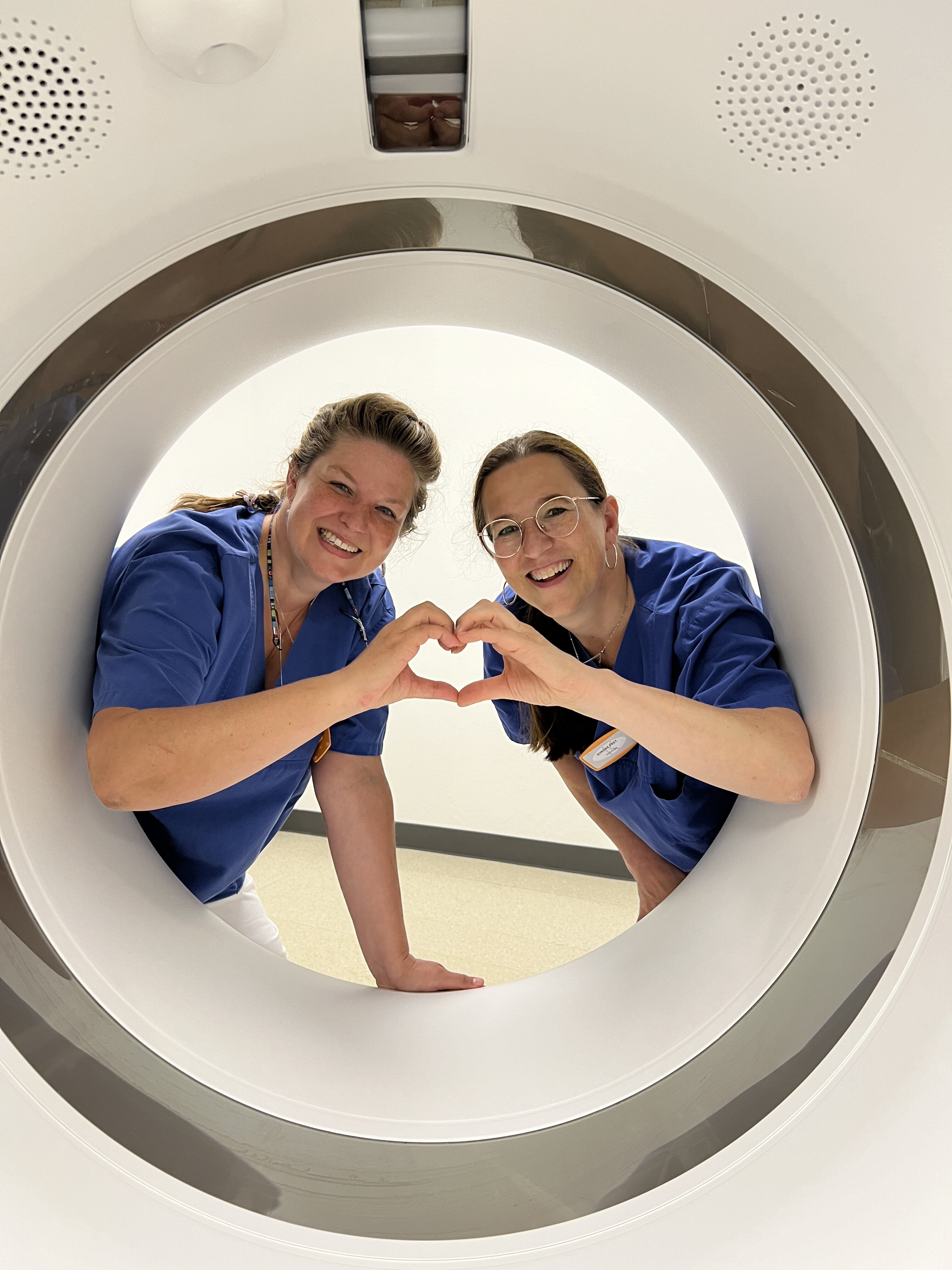 Zwei Medizinische Technologen für Radiologie schauen durch eine MRT Rähre und formen mit den Fingern ein gemeinsames Herz. 