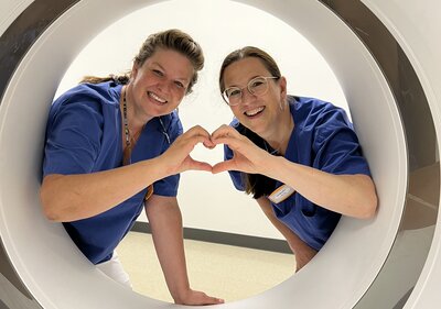 Zwei Medizinische Technologen für Radiologie schauen durch eine MRT Rähre und formen mit den Fingern ein gemeinsames Herz. 