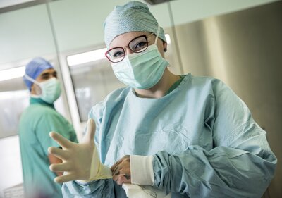ine Person in OP-Kleidung steht in einem Operationssaal. Sie trägt einen Mund-Nasen-Schutz, eine OP-Haube und medizinische Handschuhe. Im Hintergrund ist eine zweite Person zu sehen.